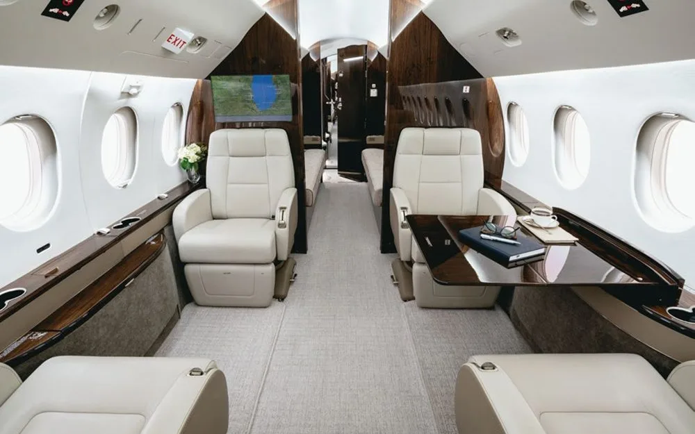 The elegant cabin interior of a Dassault Falcon 900EX, showing light leather seats, a deployed table with a book and coffee, and dark wood paneling.