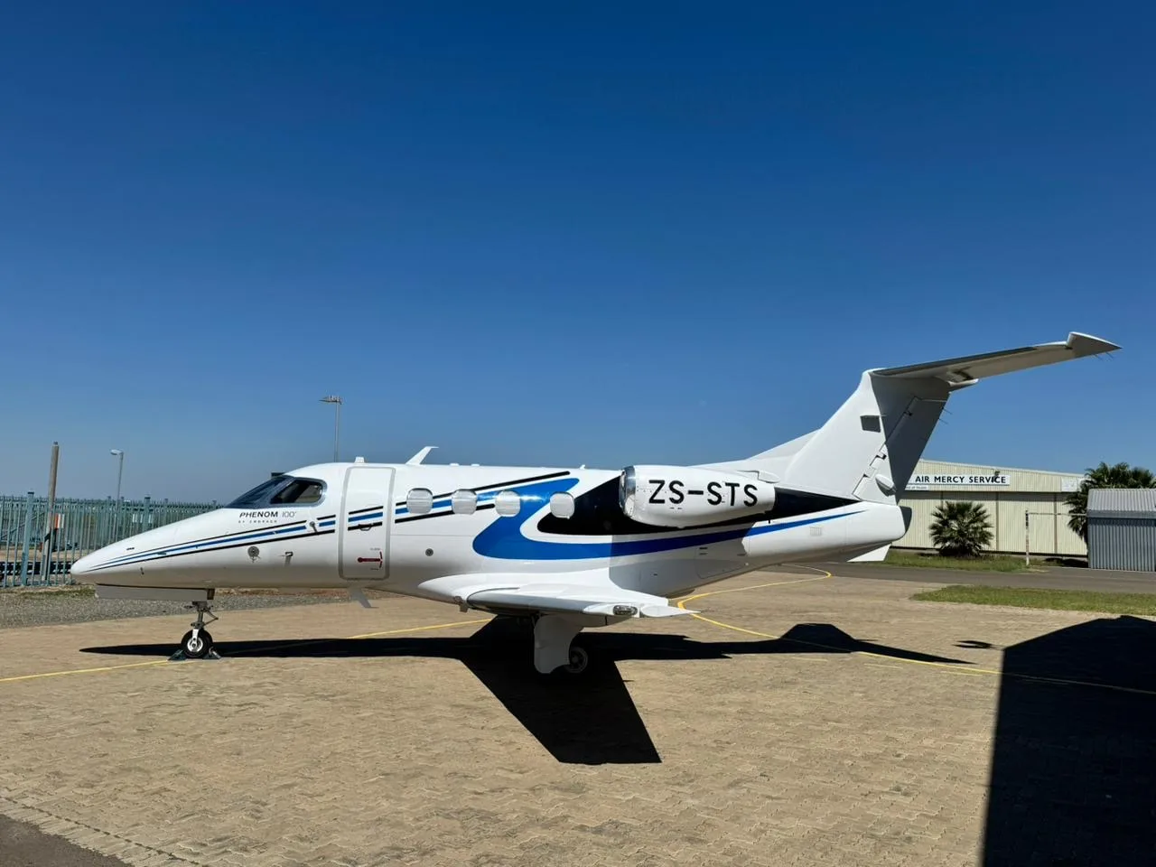 A white Embraer Phenom 100 business jet with blue and black stripes and registration ZS-STS is parked on an airport apron on a sunny day.