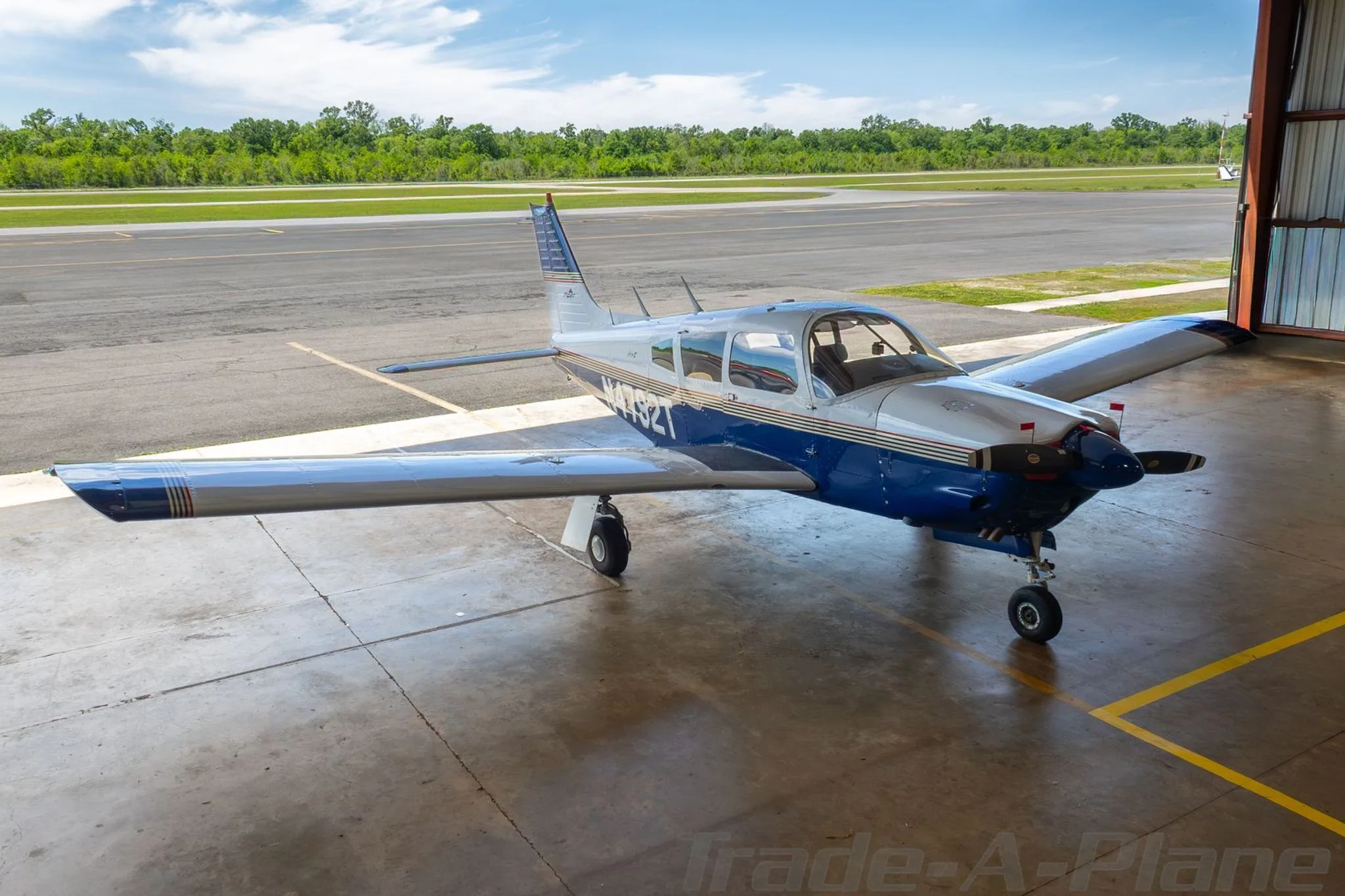 A blue and white Piper Arrow II aircraft, registration N4792T, parked on the tarmac outside a hangar.