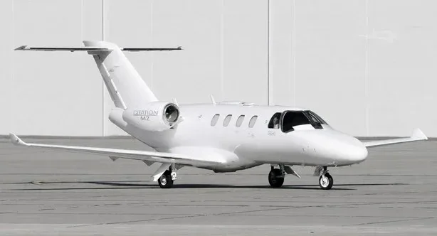 A white Cessna Citation M2 business jet is parked on an asphalt tarmac with a white building in the background.
