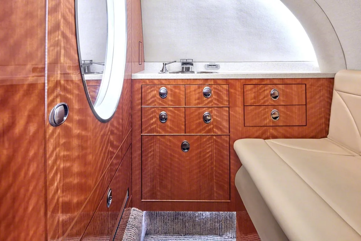 A close-up view of the aircraft interior showing polished wood cabinetry, a small sink, and a beige seat.