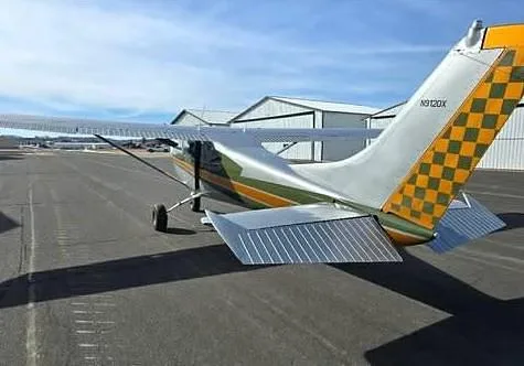 The tail section of a Cessna 182D aircraft with N9120X registration and a yellow and black checkered pattern, parked on an airfield tarmac.
