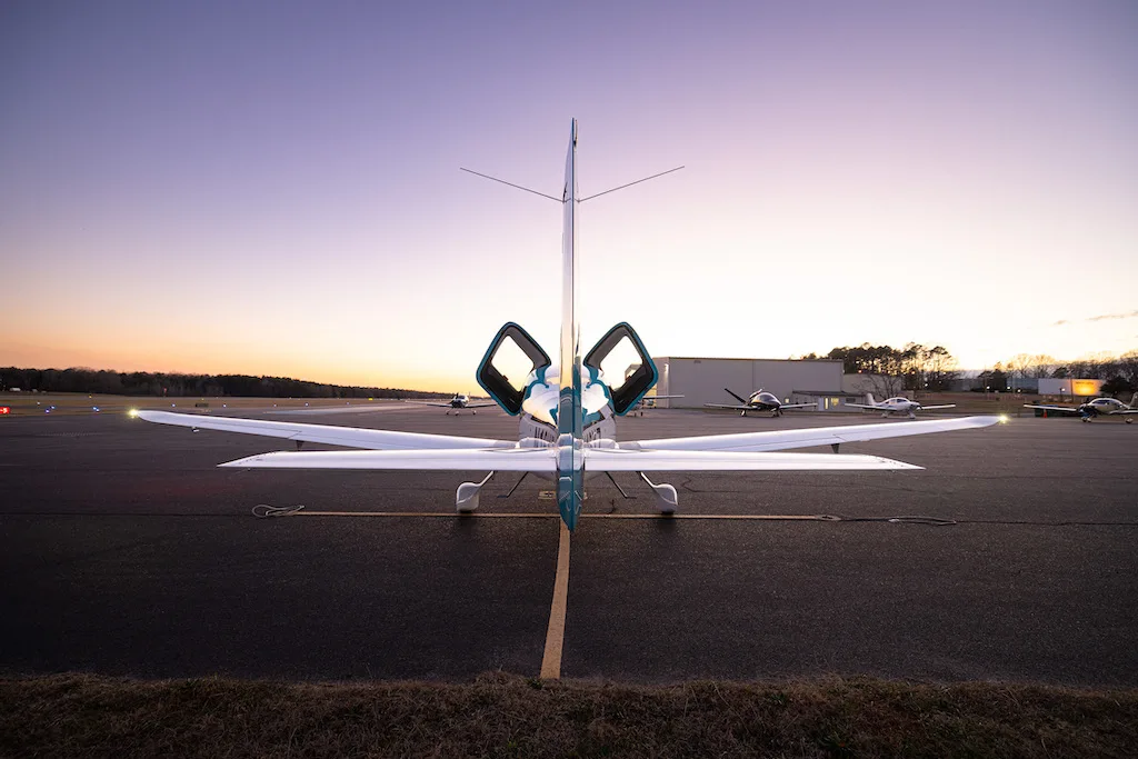 A rear view of a turquoise and white Cirrus SR22 aircraft with its doors open, parked on an airfield at dusk.