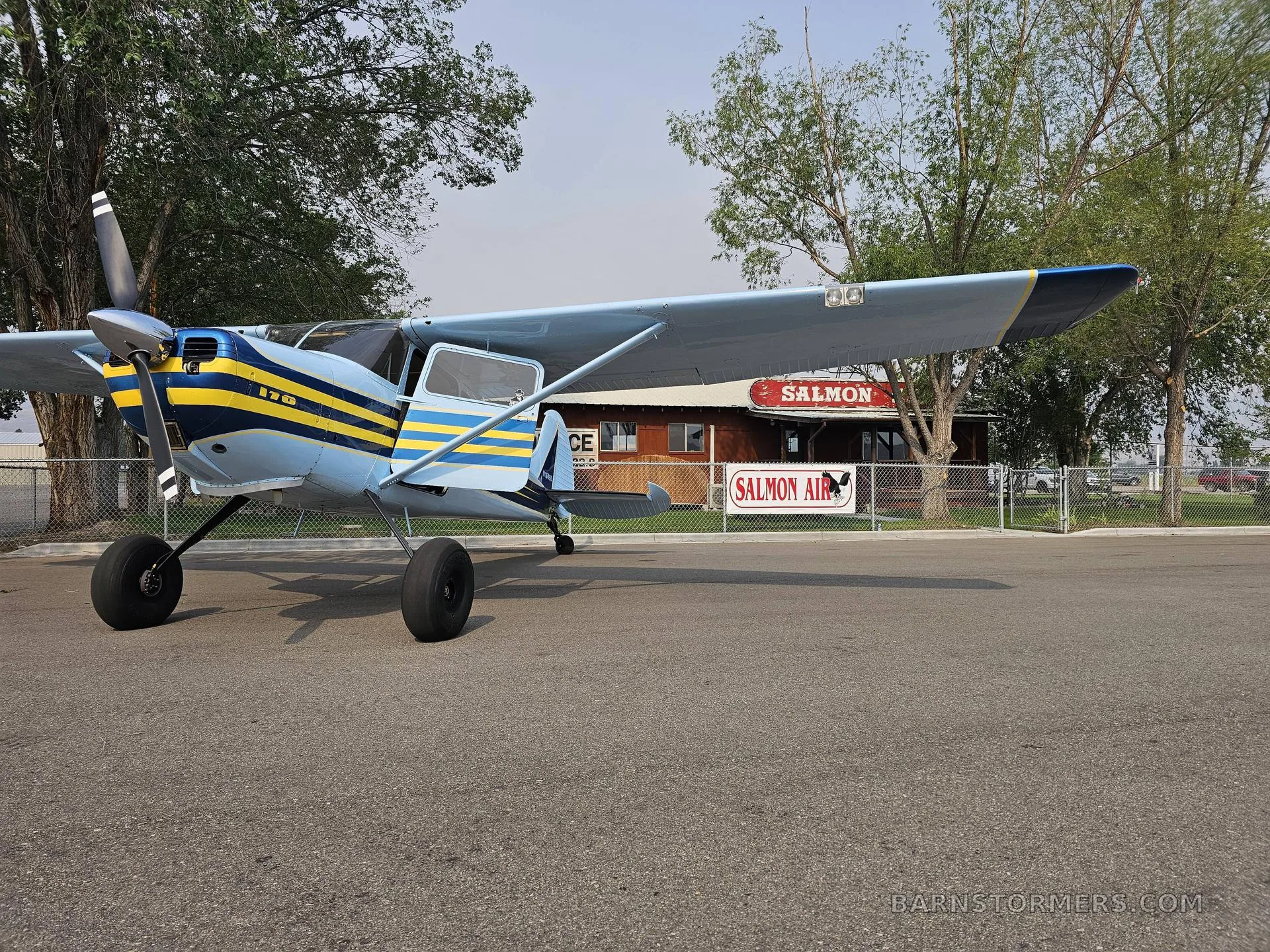 A light blue and yellow striped Cessna 170 aircraft is parked on an asphalt apron in front of a "SALMON AIR" building.