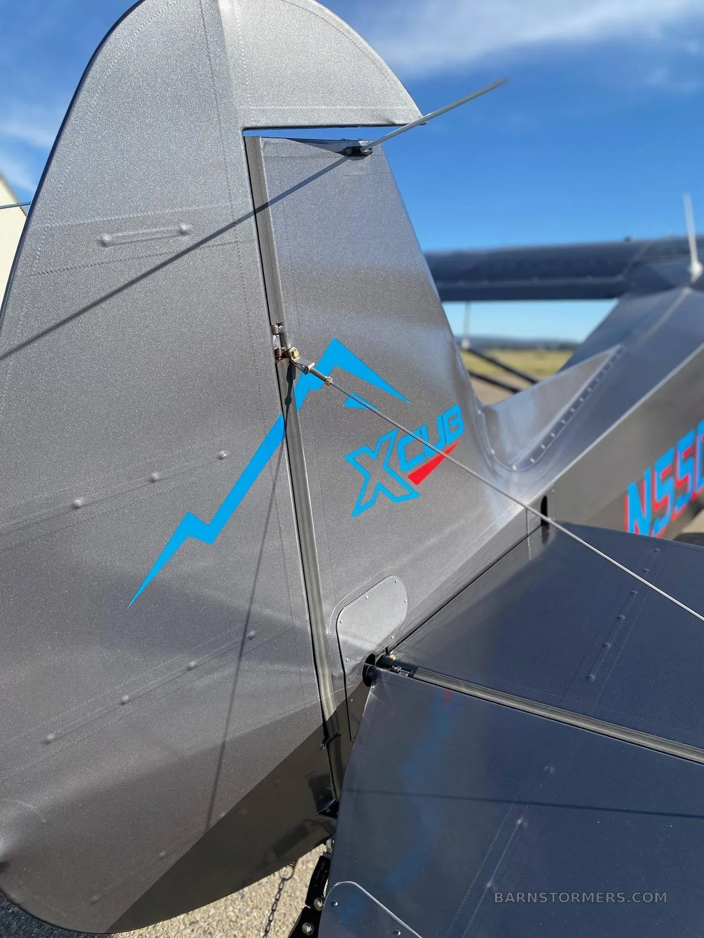 A close-up view of the tail of a dark gray X-Cub aircraft, showing the blue 'XCUB' logo and registration N55CX.