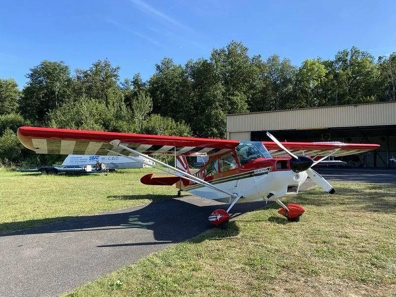 A red and white Super Decathlon aircraft (N184AC) is parked on grass next to an asphalt path and a hangar.