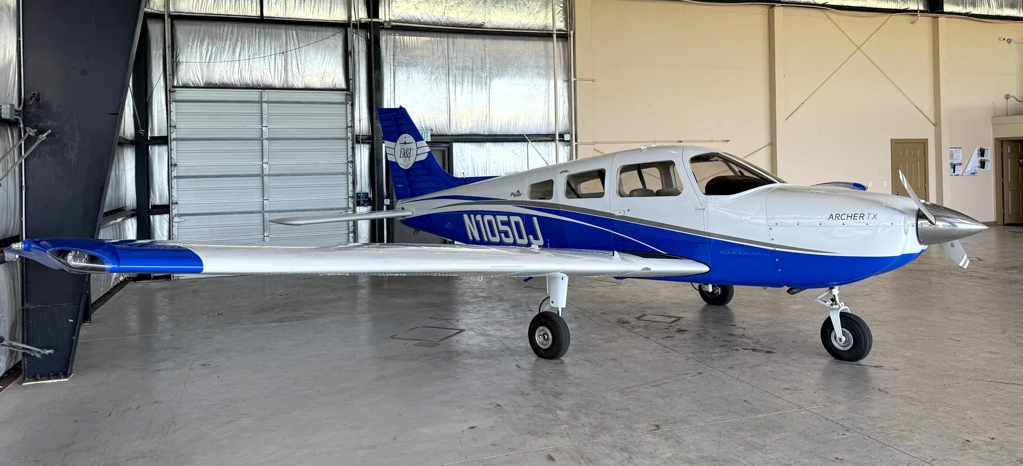 A white and blue Piper Archer TX aircraft, N105DJ, is parked inside a hangar.