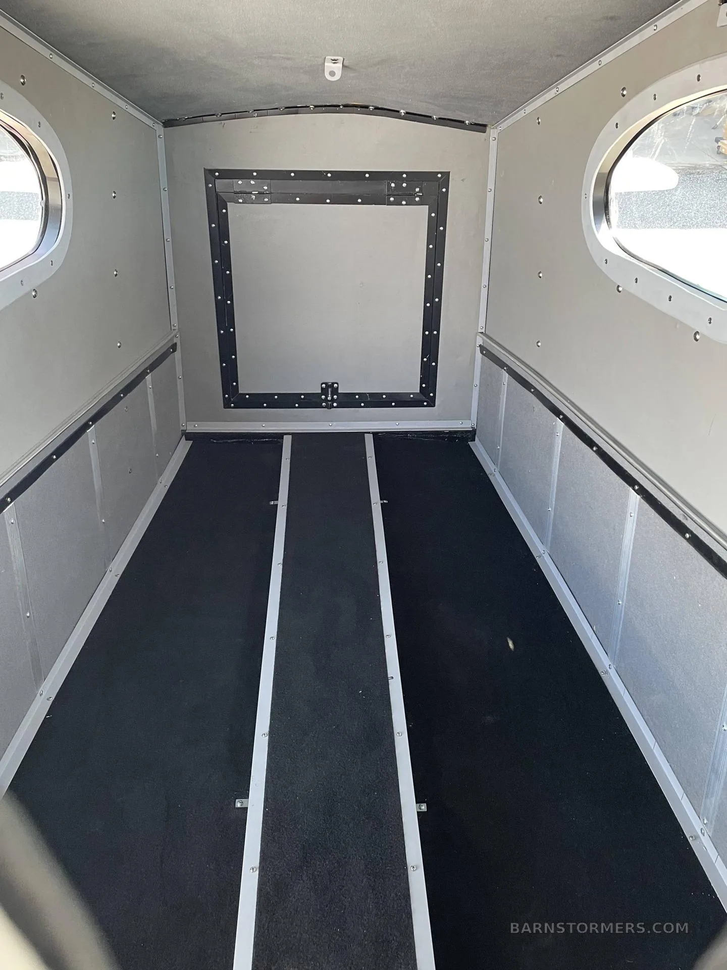 The empty cargo area of an aircraft features black floor mats and two oval windows.