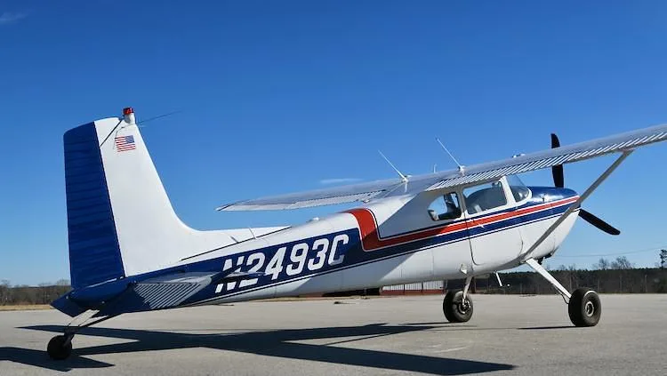 A side view of a white and blue Cessna 180 Skywagon with red stripes, registration N2493C, parked on an airfield.