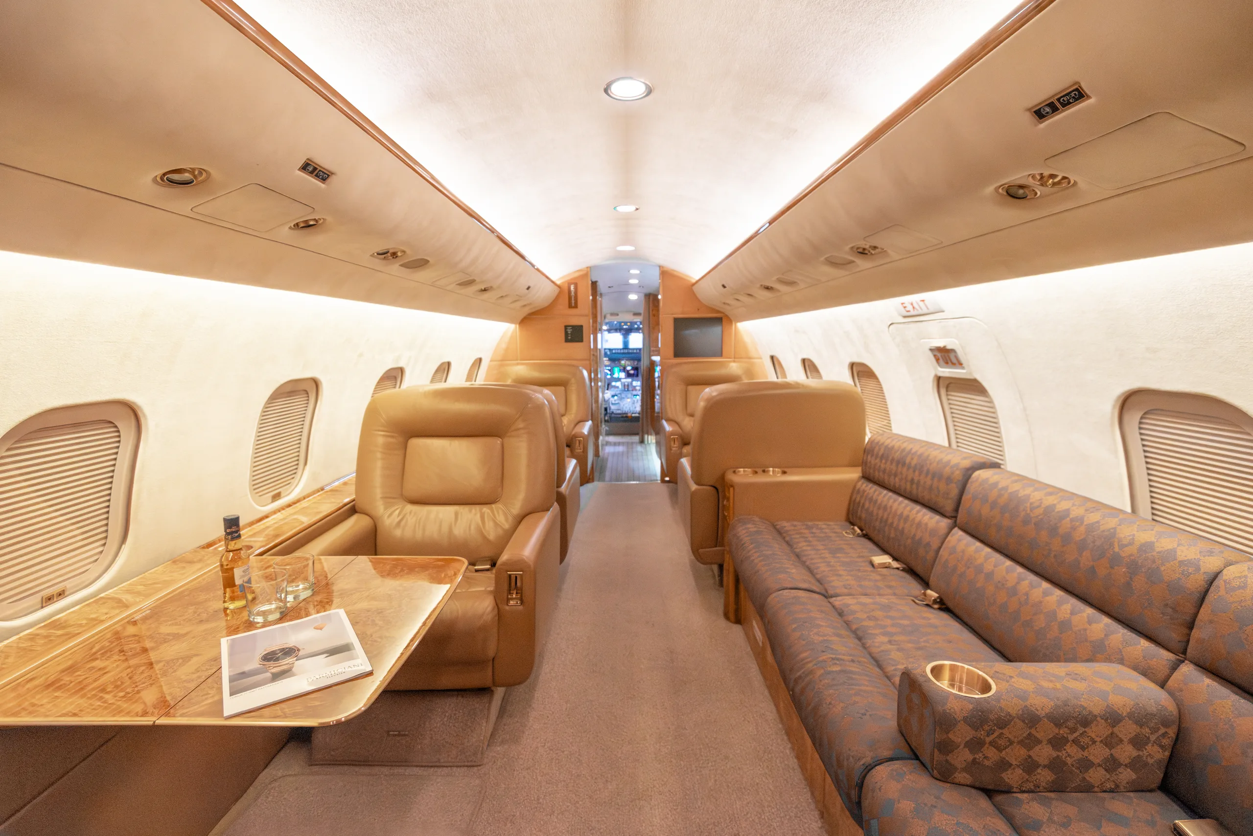 The private jet cabin interior featuring tan leather seats, a patterned sofa, and a table with drinks and a magazine.