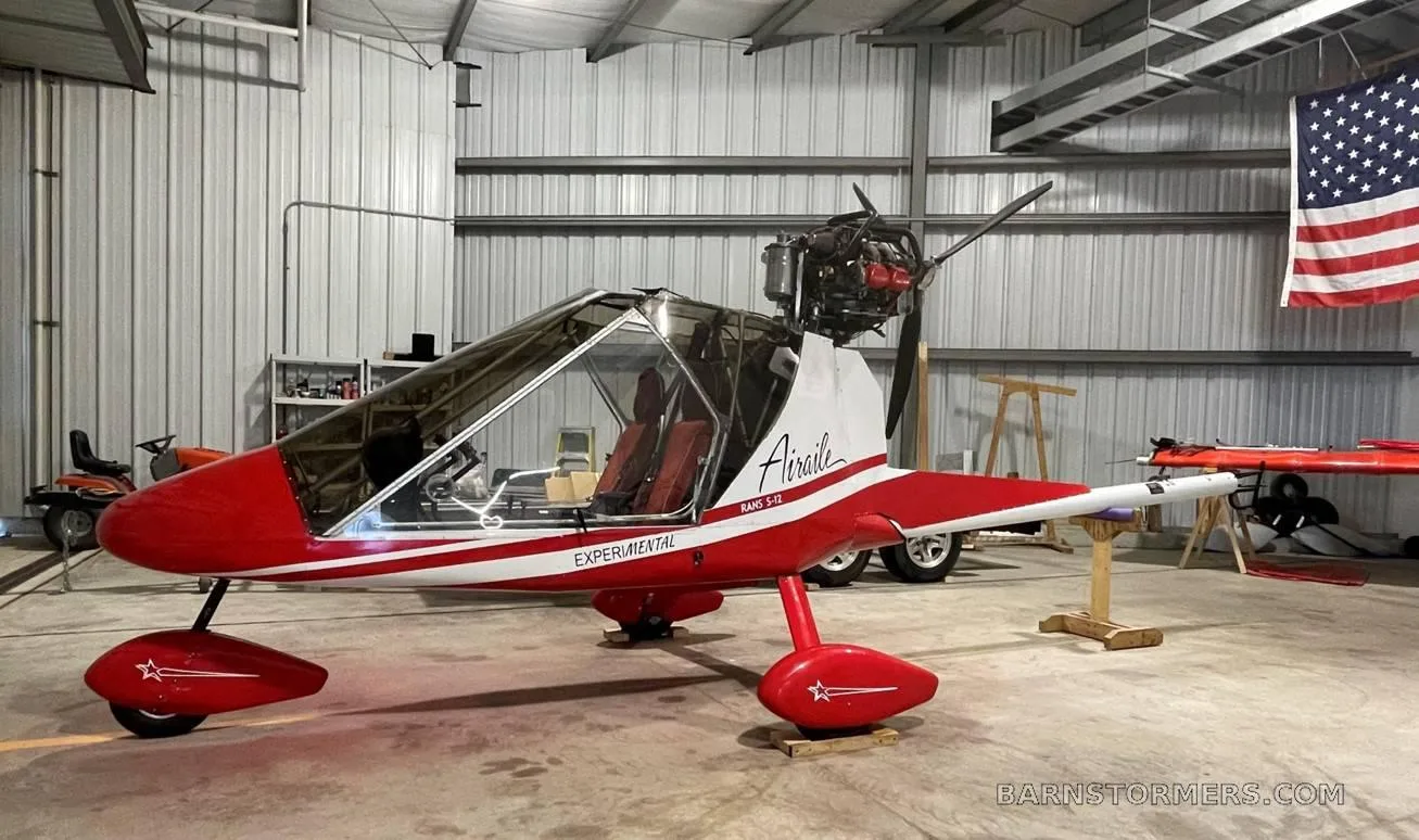 A red and white RANS S-12 Airaile experimental aircraft with a pusher propeller, parked in a hangar.