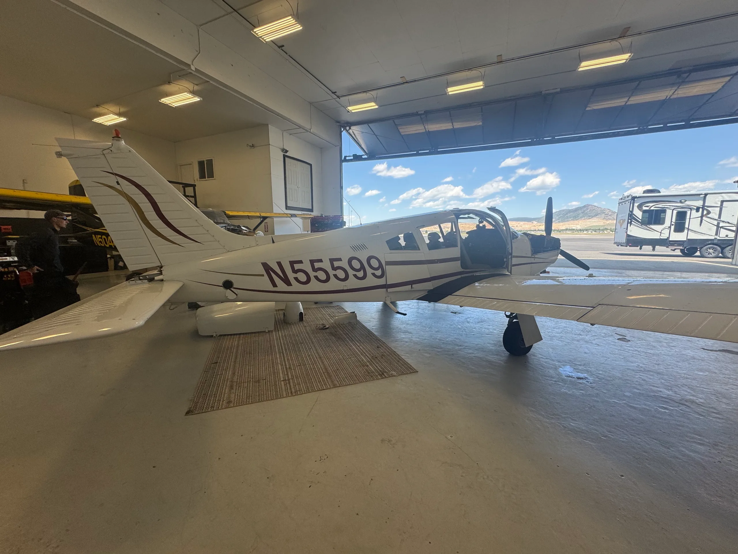 A white single-engine propeller aircraft, N55599, with maroon and gold stripes, parked inside a hangar with its door open to a sunny day.
