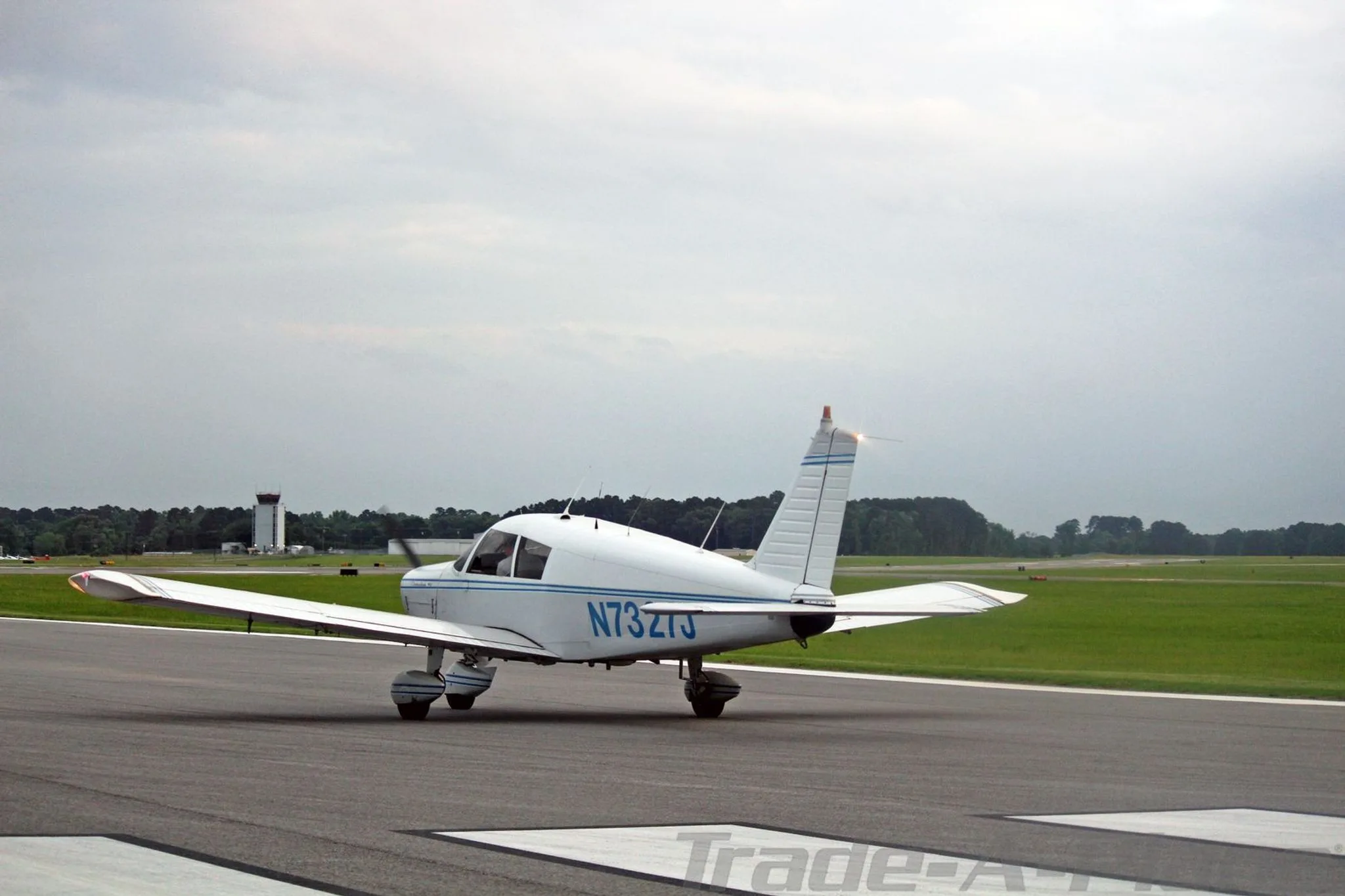 A white Piper Cherokee 140 aircraft, N7327J, with blue stripes, taxis on an airport runway with a control tower in the background.