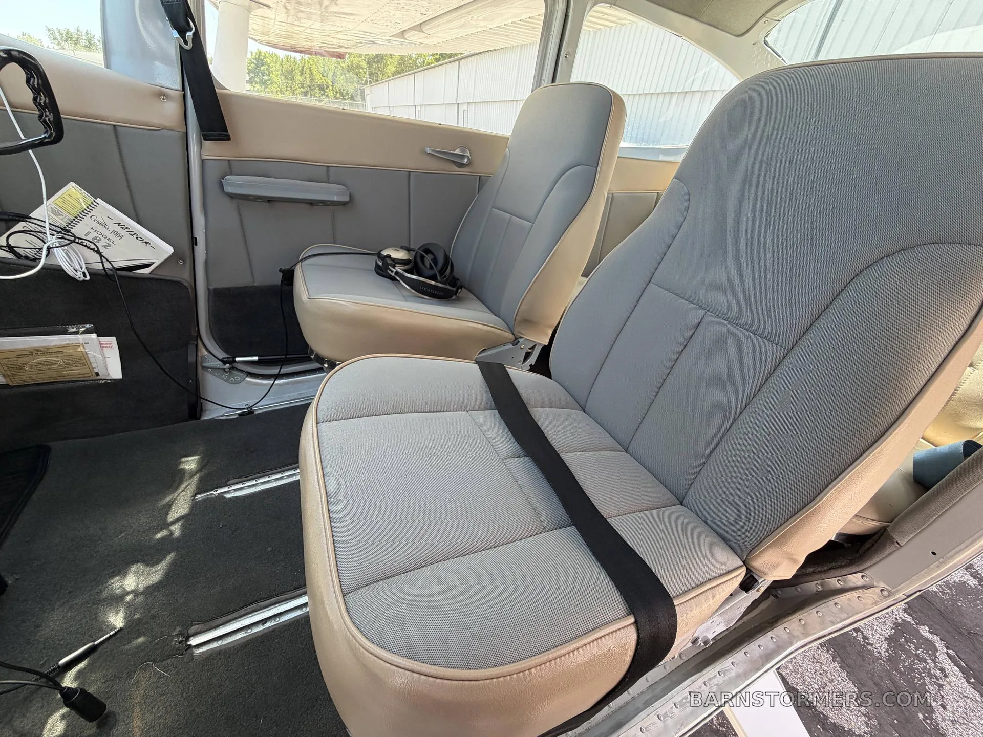 The light gray and tan interior cabin of a small aircraft, showing two front seats with seatbelts.