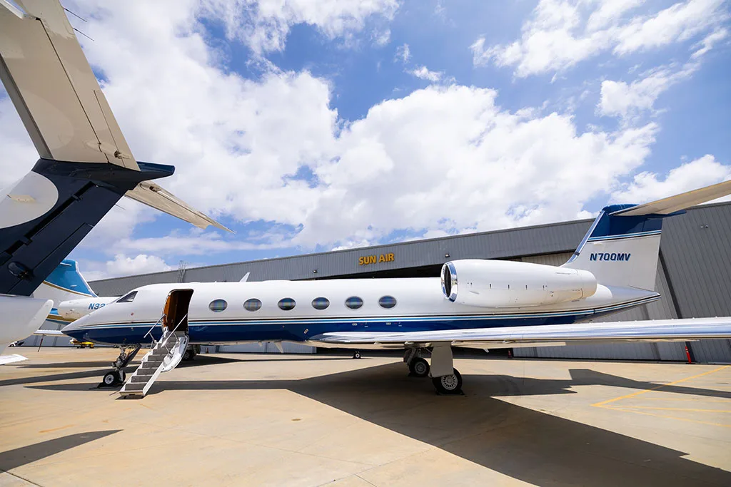 A white Gulfstream G550 with blue stripes, tail number N700MV, parked on an tarmac with stairs deployed, in front of a hangar.