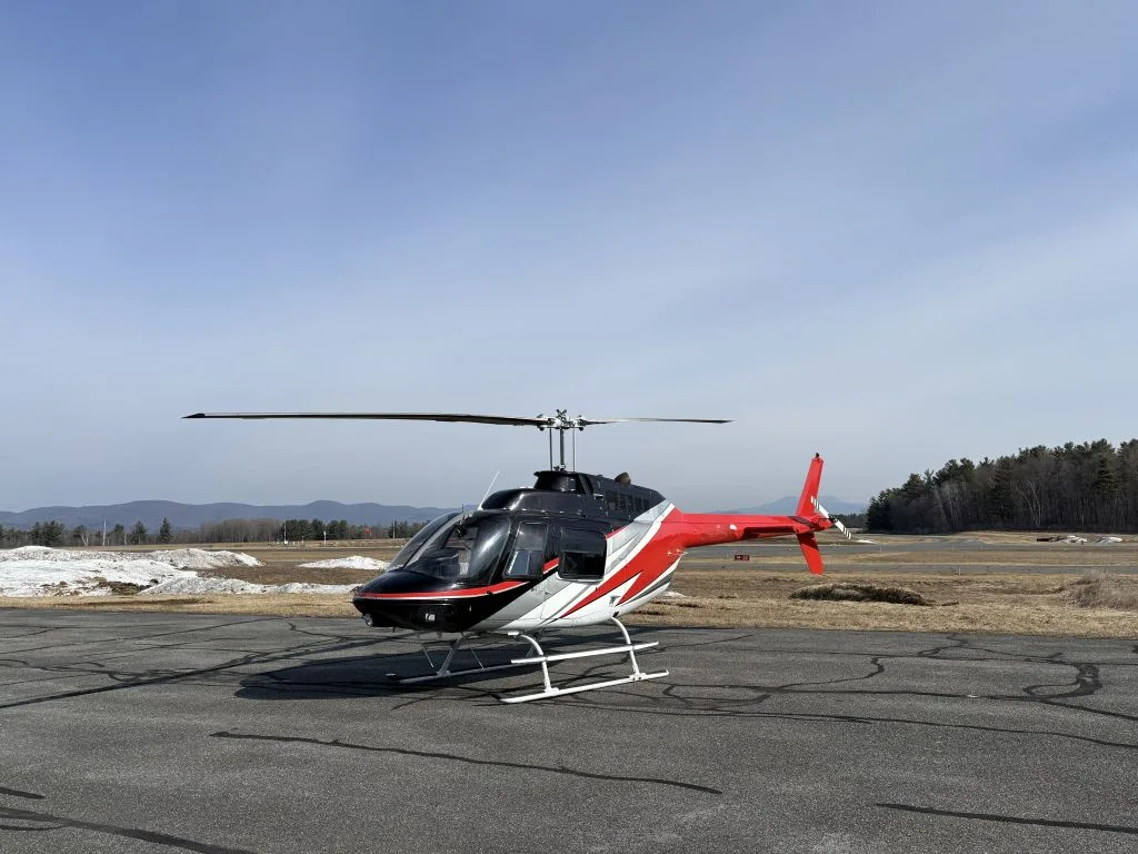 A black, white, and red helicopter is parked on an asphalt surface with a snowy landscape and trees in the background.
