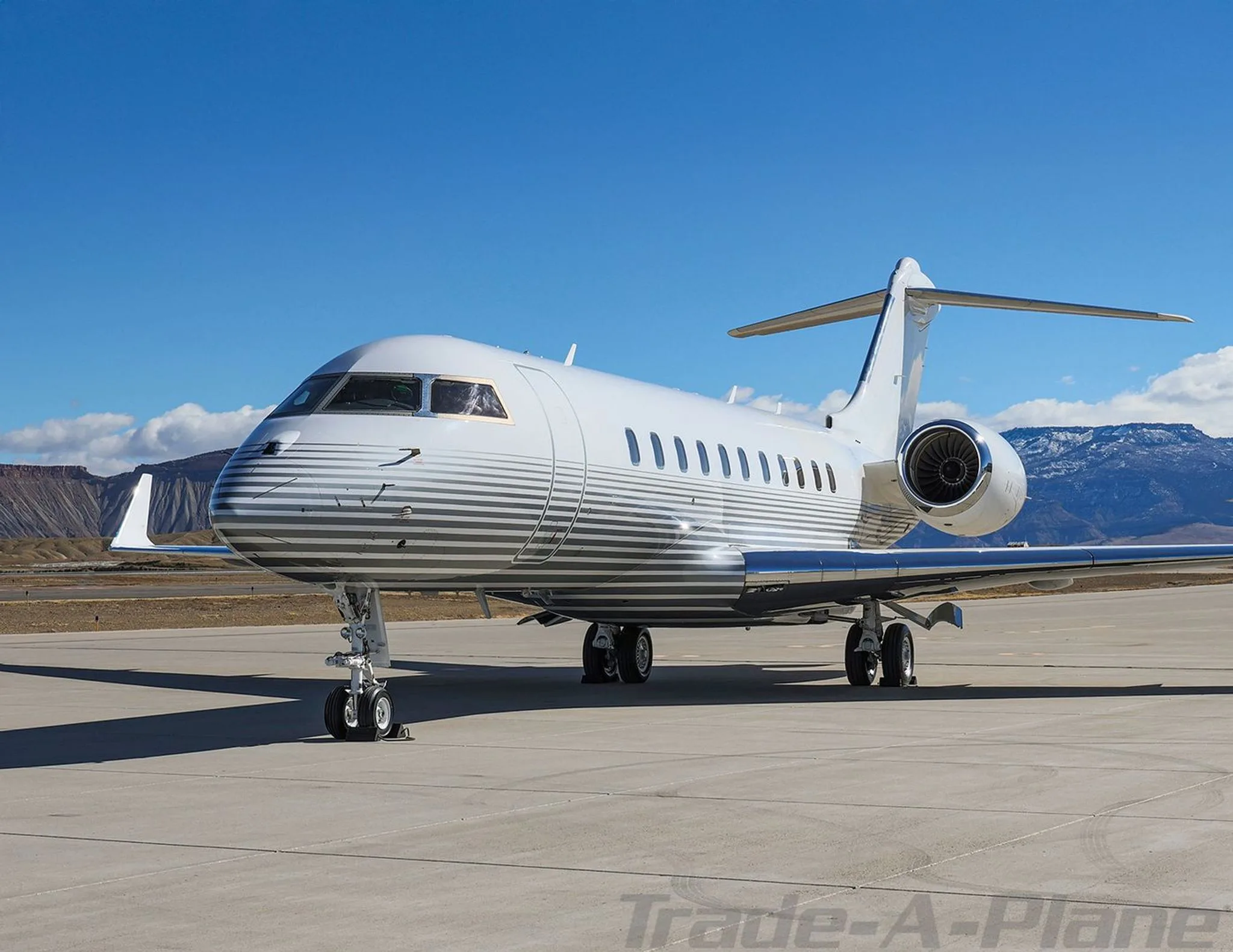 A front-side view of a white Bombardier Global 6000 business jet with grey pinstripes on the fuselage, parked on a tarmac.