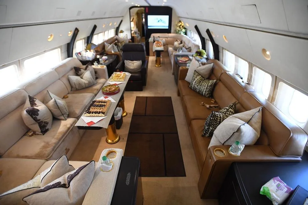 A wide view of a spacious aircraft cabin with multiple seating areas, including sofas and armchairs, and a large screen at the far end.