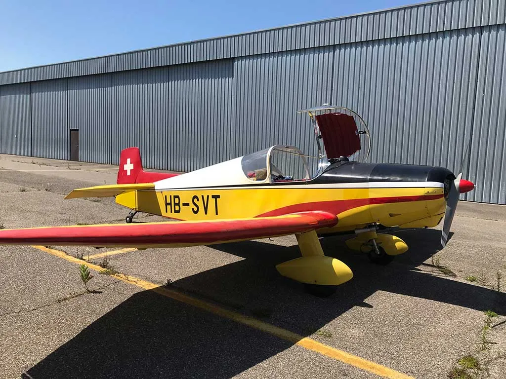 A yellow, red, and black single-engine aircraft (HB-SVT) with its canopy open, parked on an asphalt tarmac next to a hangar.