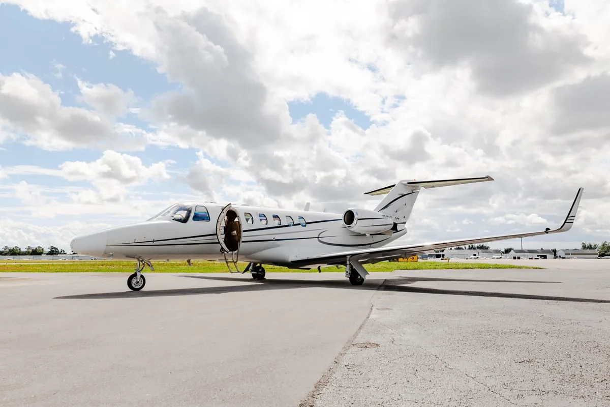 A white business jet with a black stripe sits on an airport tarmac under a partly cloudy sky with its entry door open.