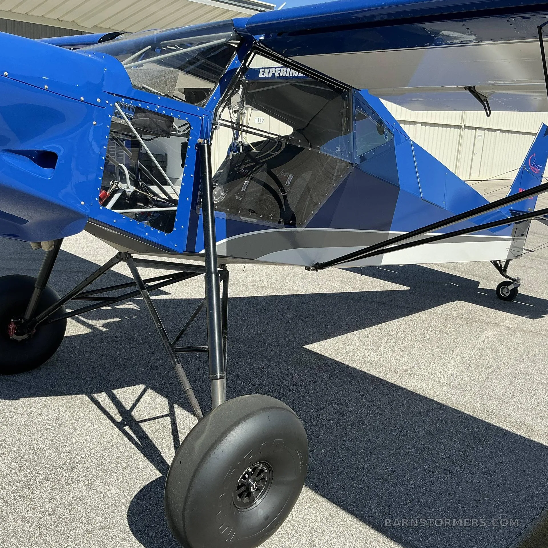 A side view of the blue and white SuperSTOL aircraft's fuselage, landing gear with large tires, and open cabin door.