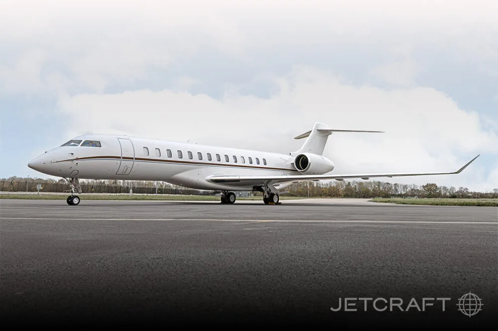 A white Bombardier Global business jet with brown accent stripes parked on an airfield under a cloudy sky.