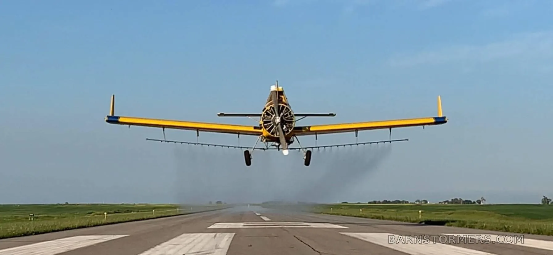 A yellow agricultural aircraft with blue stripes taking off from a runway, viewed from behind, with spray visible.