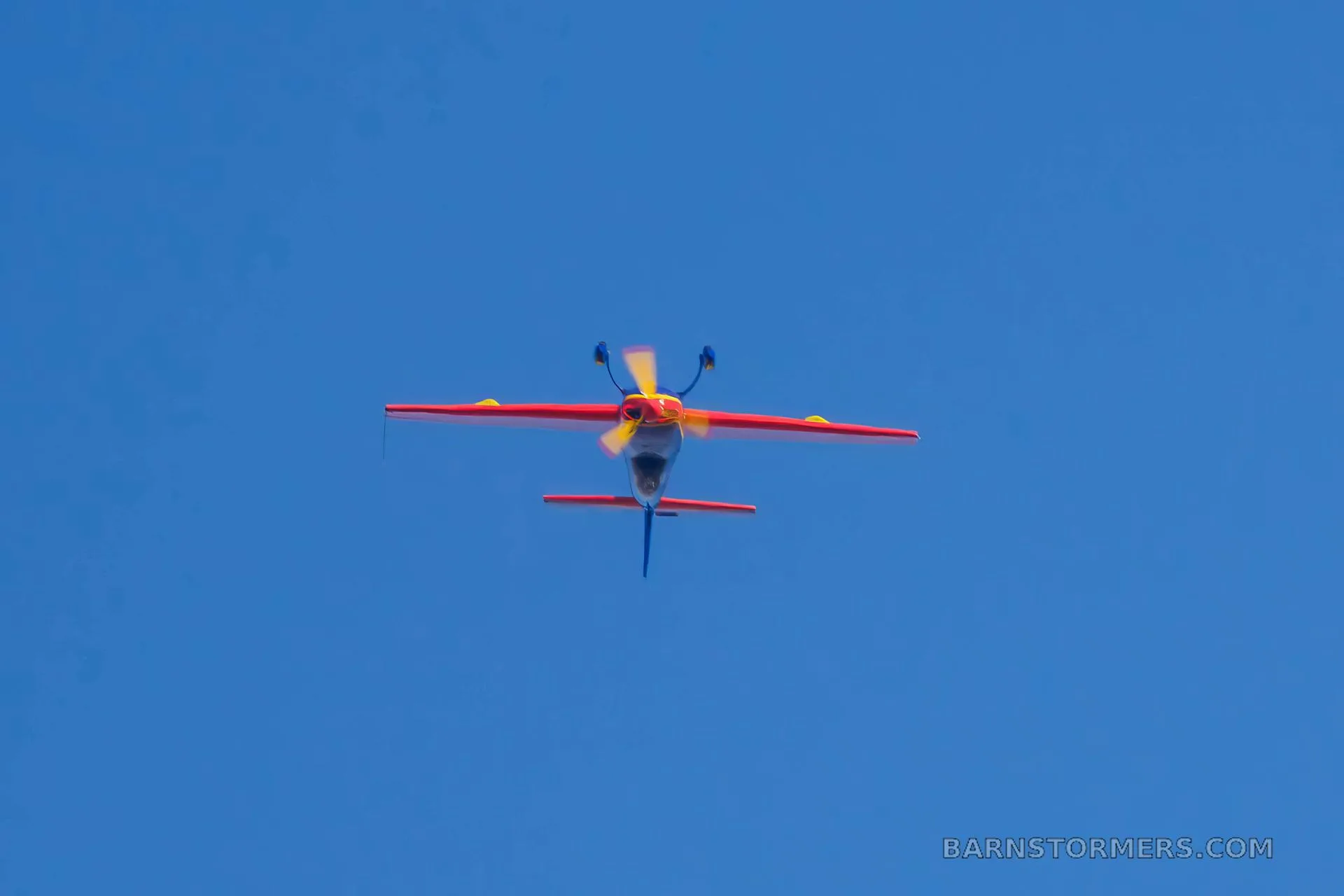 A red and blue aerobatic aircraft is seen flying head-on against a clear blue sky.