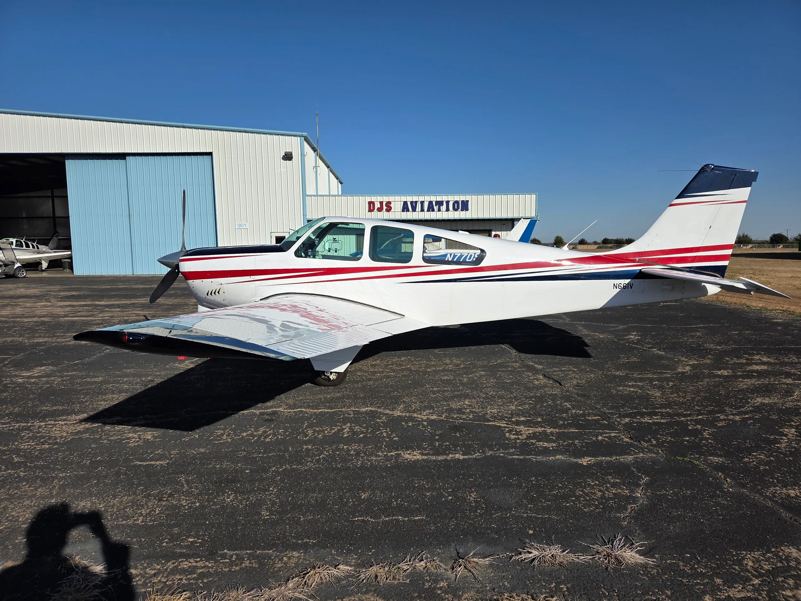 A white and red single-engine aircraft with tail number N661V parked on an asphalt ramp next to a hangar.