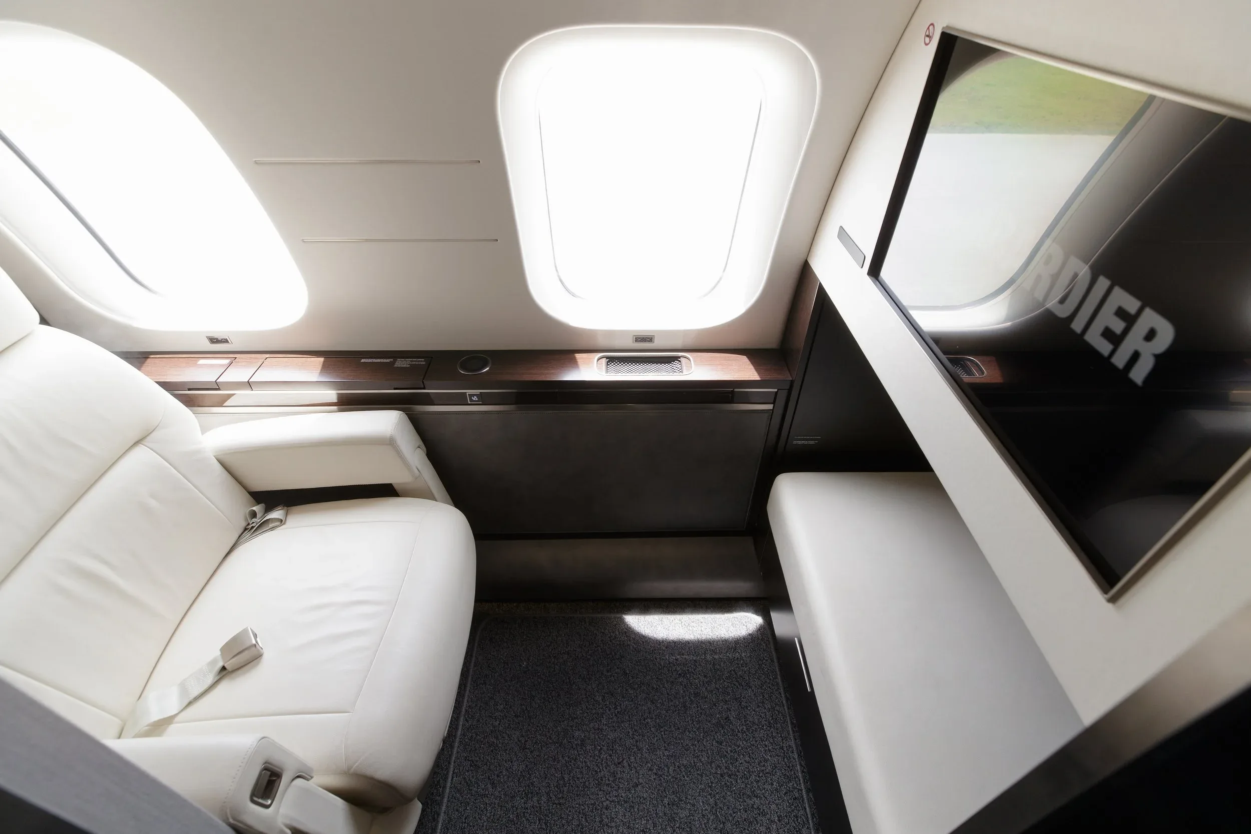 A luxurious white leather seat next to a window in a private jet cabin, with a screen displaying 'BOMBARDIER'.