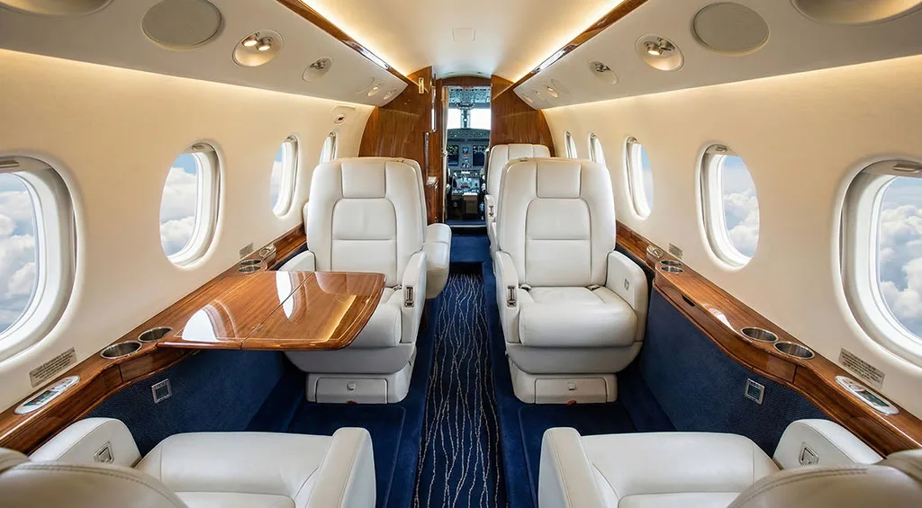 The luxurious interior cabin of a private jet features white leather seats, wooden tables, and a blue carpet, looking towards the cockpit.