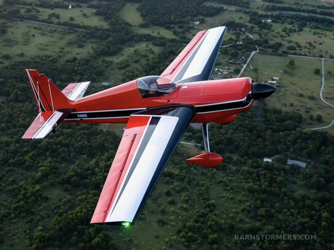 A red and white aerobatic aircraft, N300CB, with a pilot, flying over a lush green landscape.