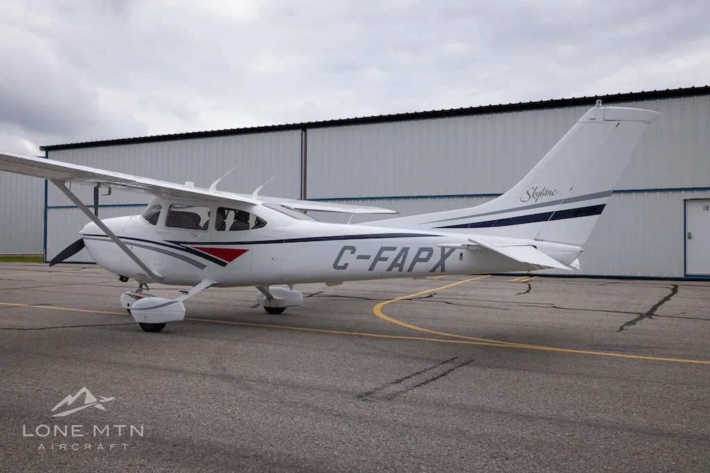 A white Cessna 182 Skylane with grey and blue stripes, registration C-FAPX, parked on an asphalt ramp with hangars in the background.