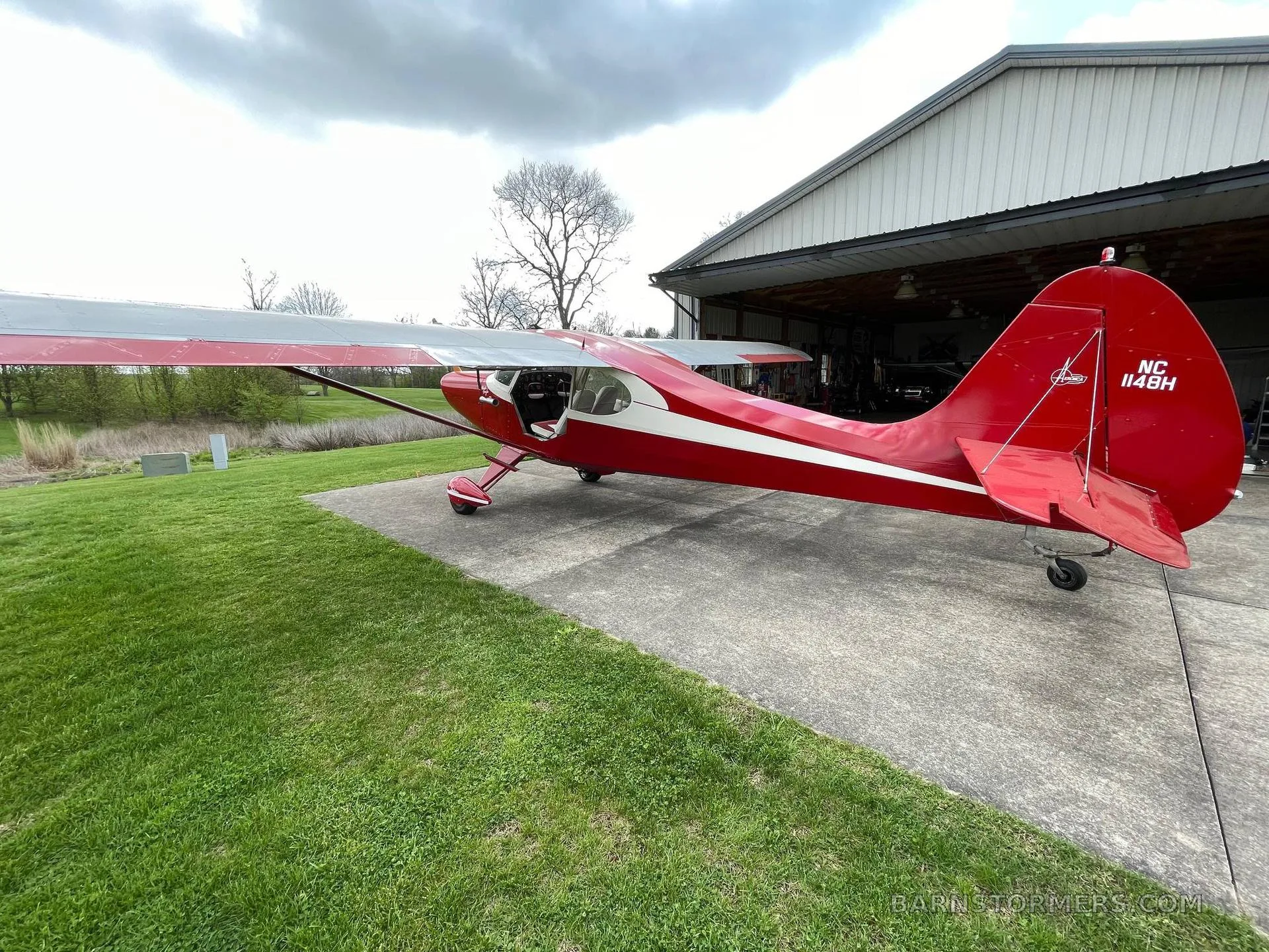 The rear-right view of a red and white single-engine propeller aircraft with registration NC 114BH, parked on concrete near a hangar.