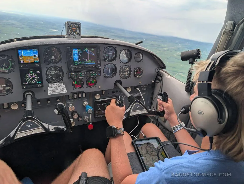 A person wearing headphones and holding the controls in the cockpit of a small aircraft, with the instrument panel and a landscape view visible.