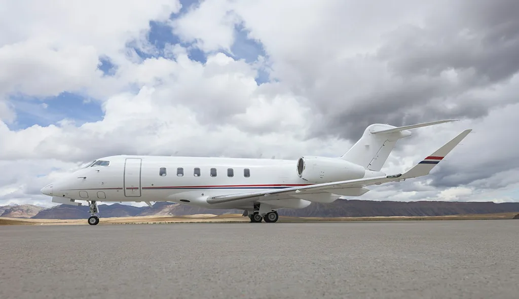 A white Bombardier Challenger 350 business jet with red and blue stripes on the tail, parked on an airfield under a cloudy sky.