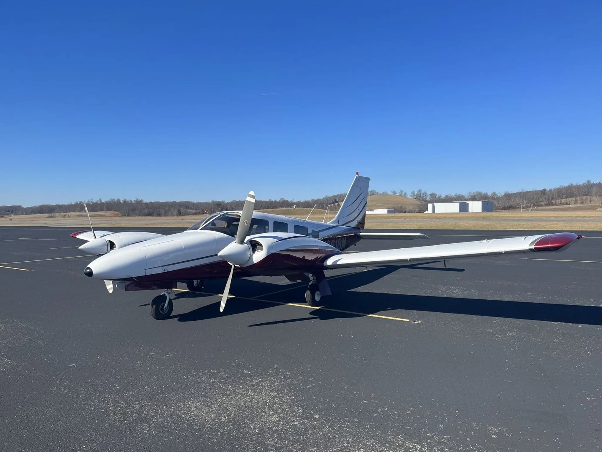 A front-side view of a white and maroon Piper N2618M twin-engine aircraft with propellers visible, parked on an asphalt tarmac.