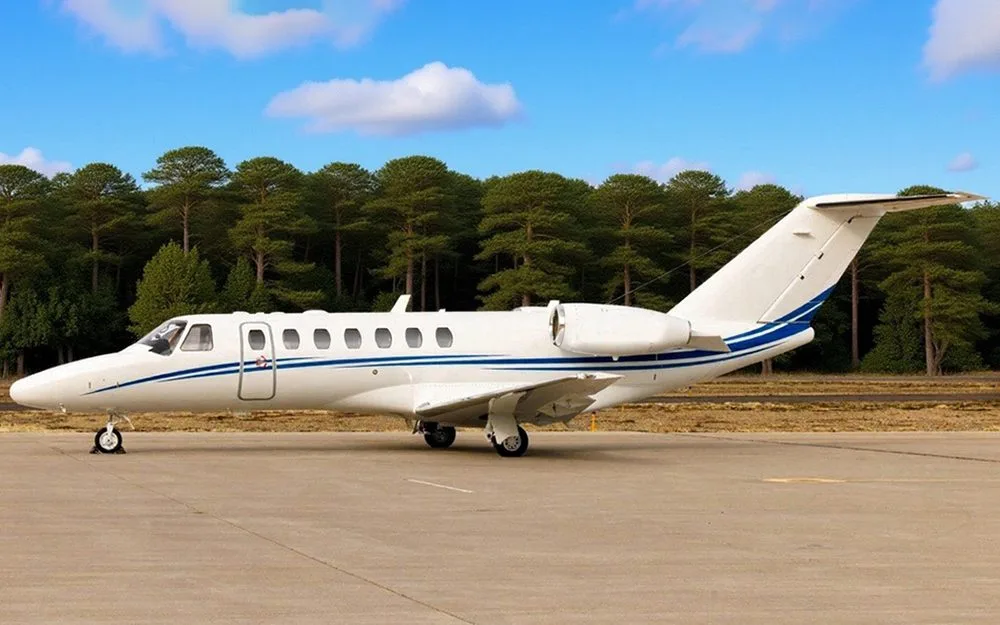 A white business jet with blue stripes on a tarmac, with a line of trees in the background under a blue sky.