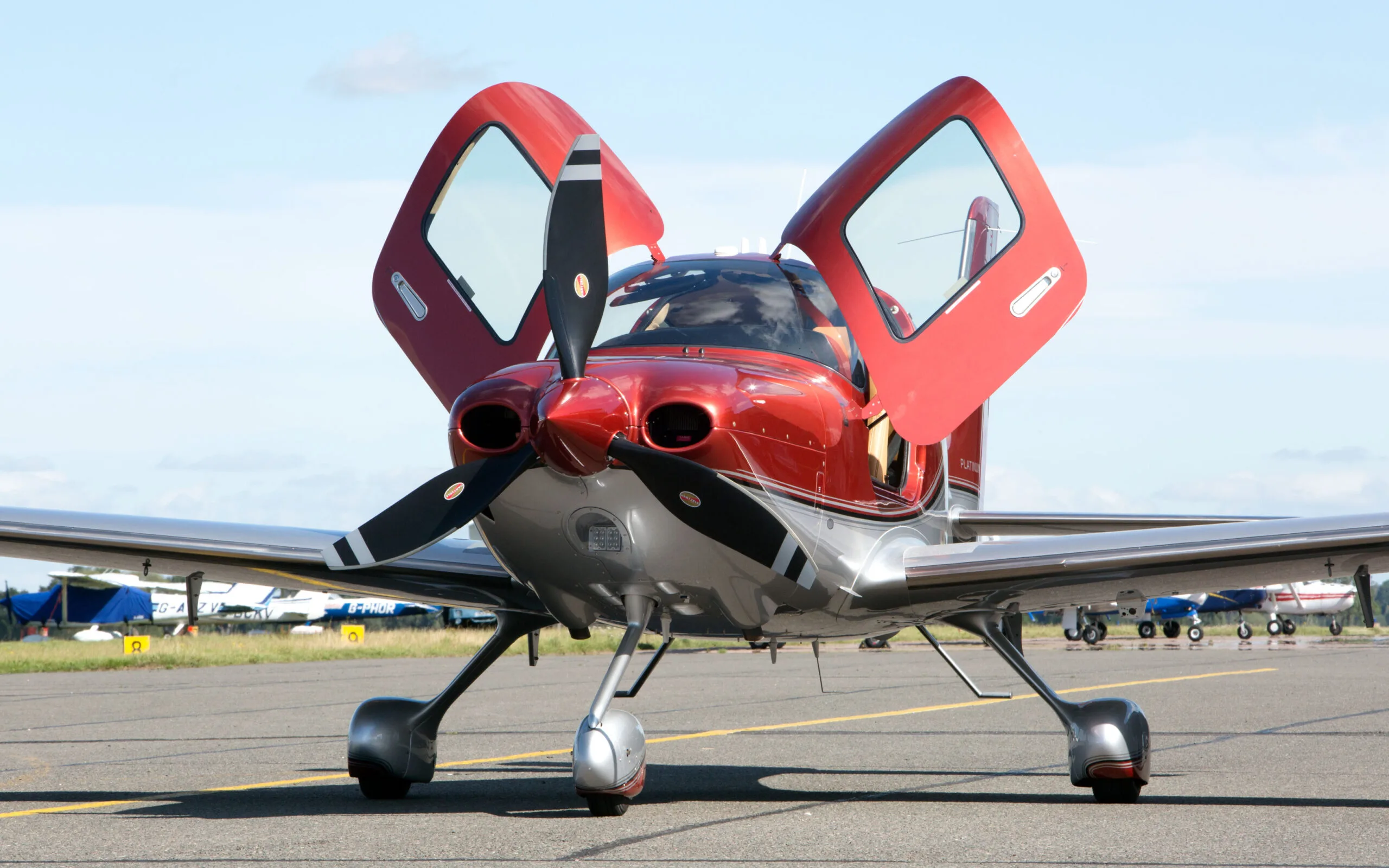 A red and silver Cirrus SR22T GTS aircraft with its gull-wing doors open, parked on an airfield tarmac under a blue sky.