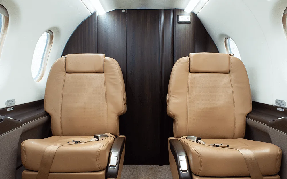 Two tan leather aircraft seats with seatbelts fastened, facing a dark wood panel wall in the cabin.