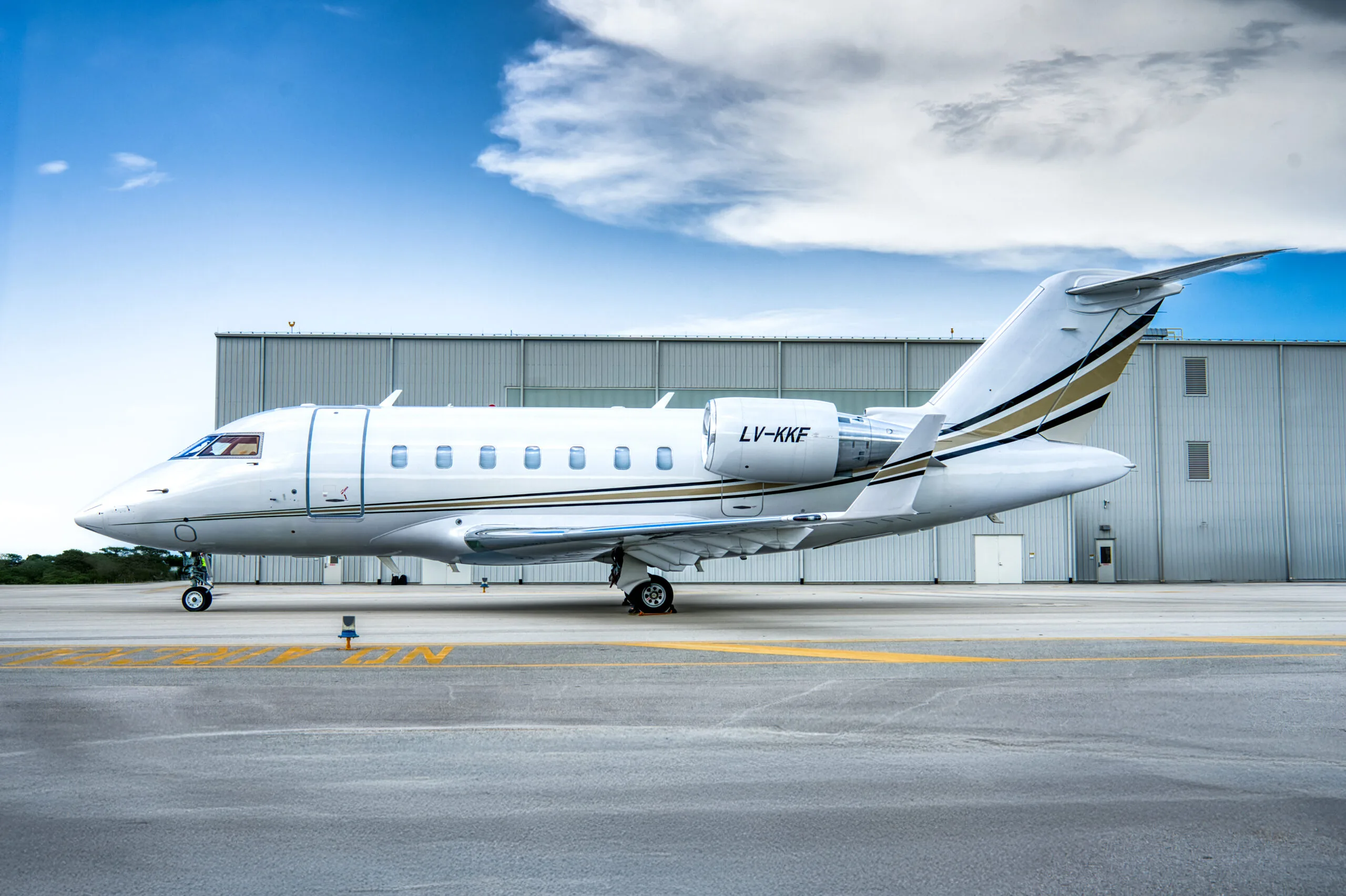A white Bombardier Challenger business jet with gold and black stripes, registration LV-KKF, seen from the side on an airport tarmac next to a hangar.