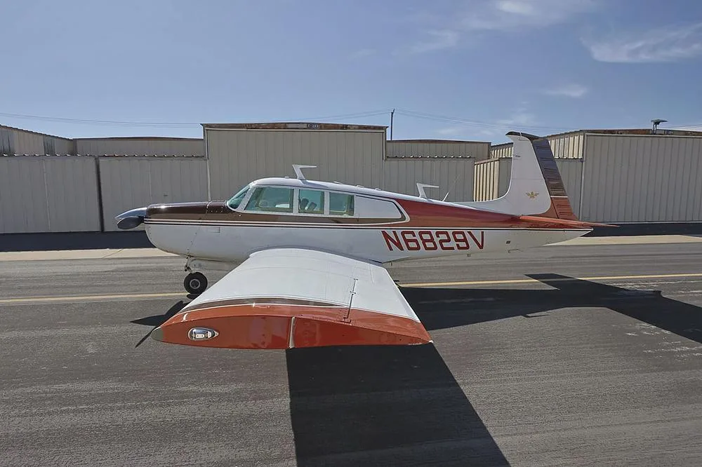 A brown and white single-engine aircraft, N6829V, is parked on an asphalt ramp with hangars in the background.