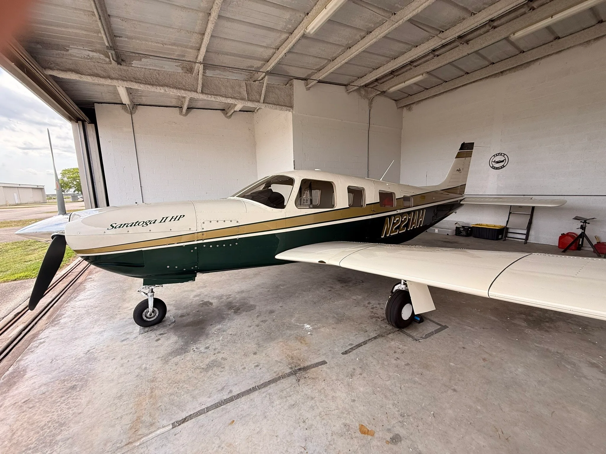 Side view of a Piper Saratoga II HP, N221AH, in a white, gold, and dark green paint scheme, parked inside a hangar.