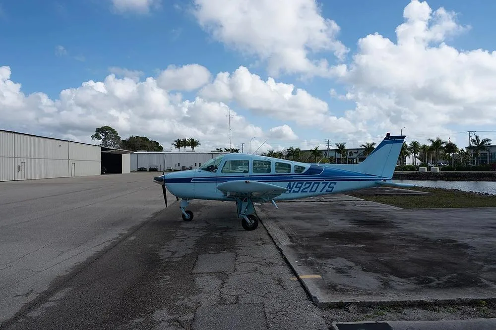 A light blue single-engine propeller aircraft, N9207S, is parked on an asphalt apron with hangars and palm trees in the background.