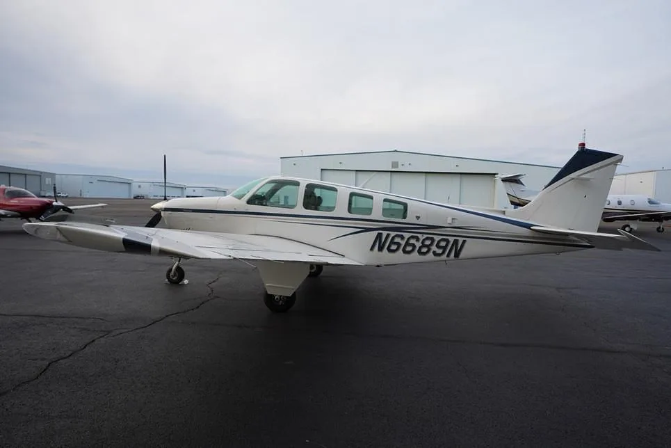 A white Beechcraft Bonanza with blue and gray stripes, registration N6689N, parked on an airport ramp with other aircraft.