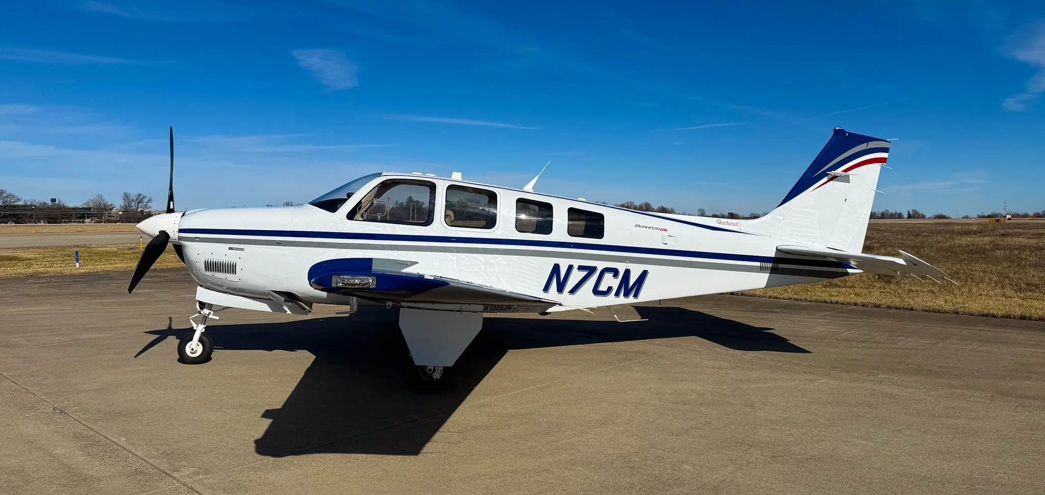 A white Beechcraft Bonanza G36 with blue and grey stripes, registration N7CM, parked on an airfield under a clear blue sky.