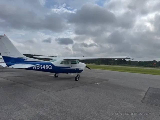 A white and blue Cessna 182 (N5146Q) is parked on an asphalt tarmac with a grassy field in the background under a cloudy sky.