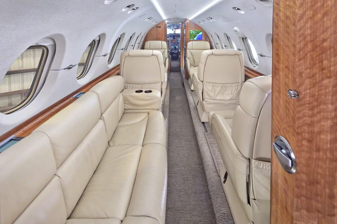 The long view of a private jet cabin with a beige leather sofa on one side and individual seats on the other.