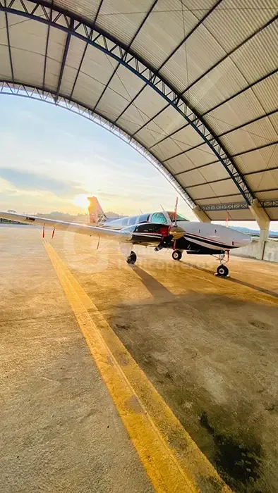 A white and black twin-engine piston aircraft parked under an open hangar structure with the setting sun in the background.