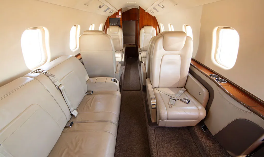 The luxurious interior cabin of a private jet with light beige leather seats and wood trim, looking towards the rear.